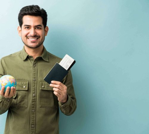 happy-young-man-holding-passport-boarding-pass-with-globe-while-standing-against-blue-background (1)